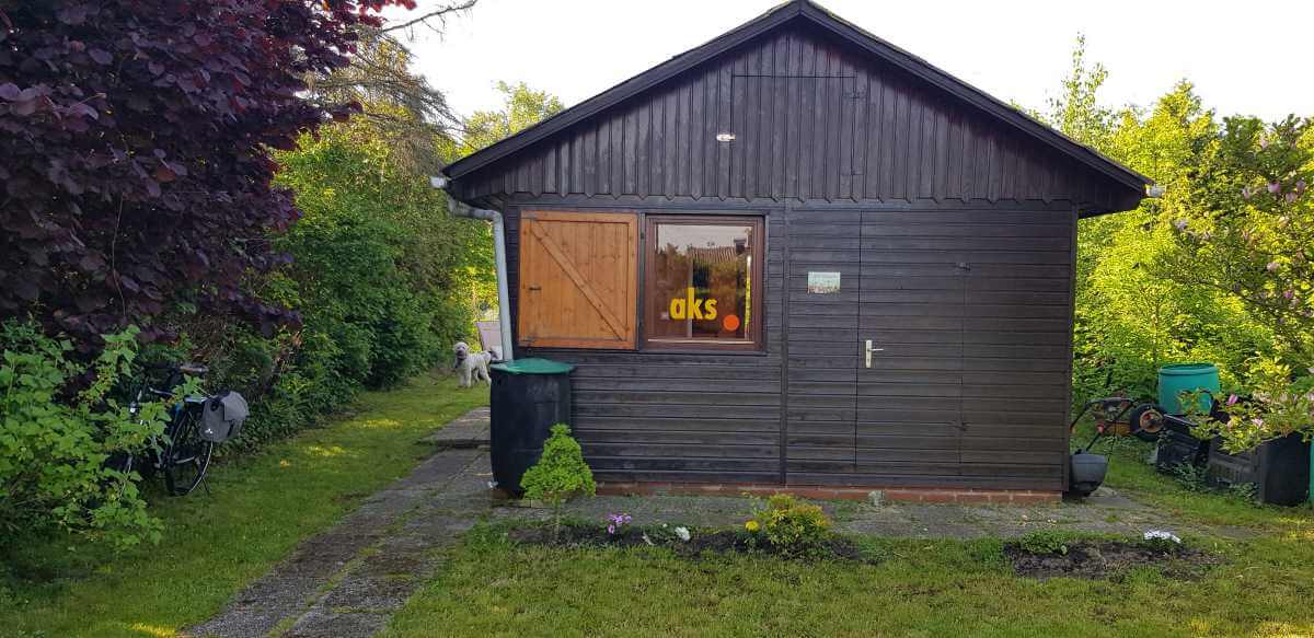 Wooden house with brown door and window with yellow lettering “aks” surrounded by green garden and bushes