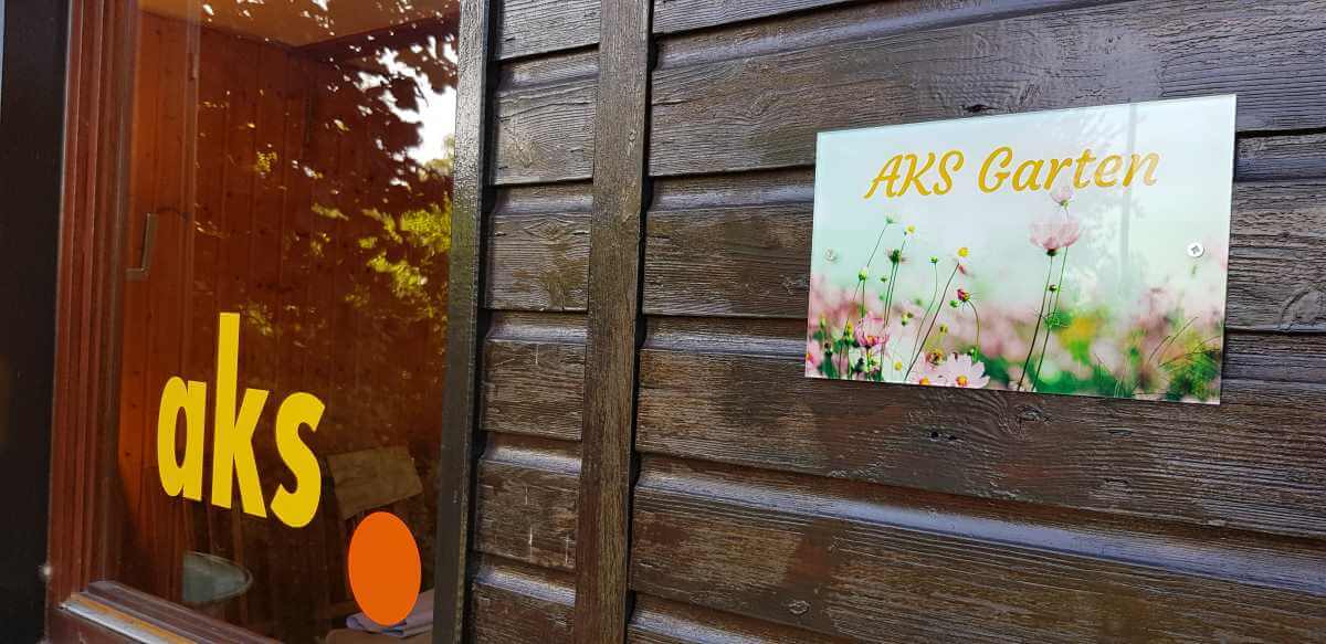 Wooden door with yellow lettering “aks” and an orange dot, as well as a sign with the inscription “AKS Garden” in front of a background with flowers
