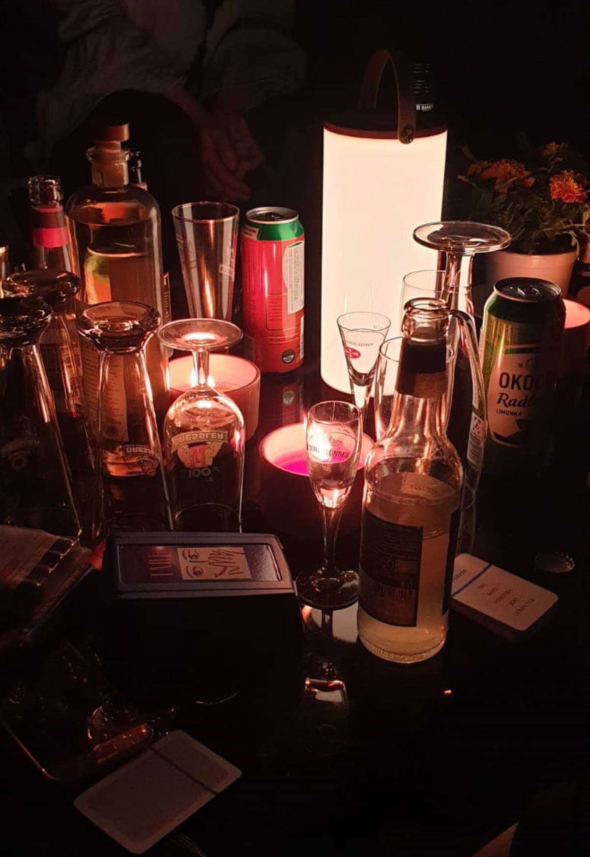 Table with various beverage bottles, cans, glasses, and an illuminated lantern at a garden party at night
