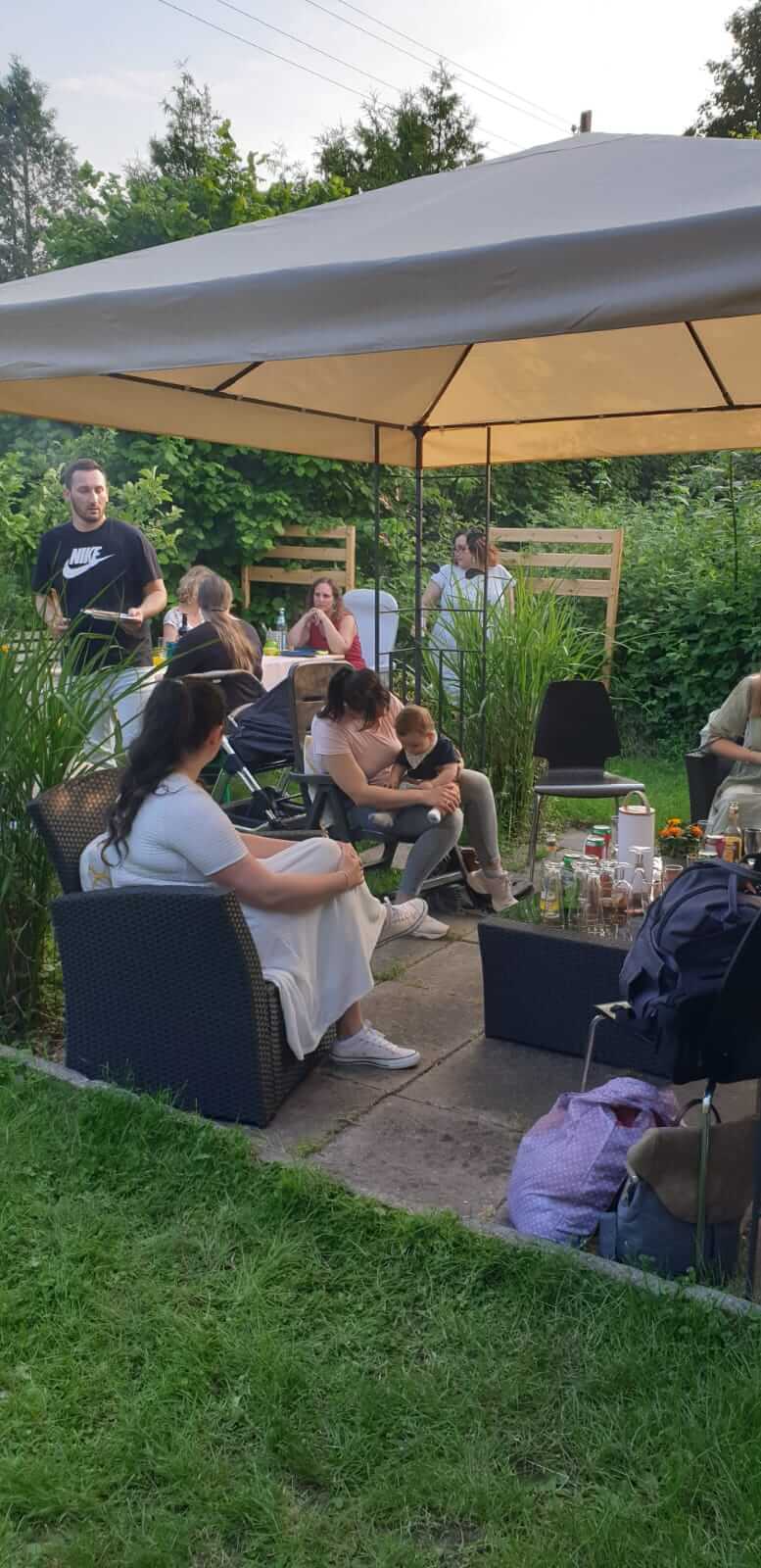 Several people are sitting and standing under a gazebo in the garden, surrounded by plants and garden furniture.