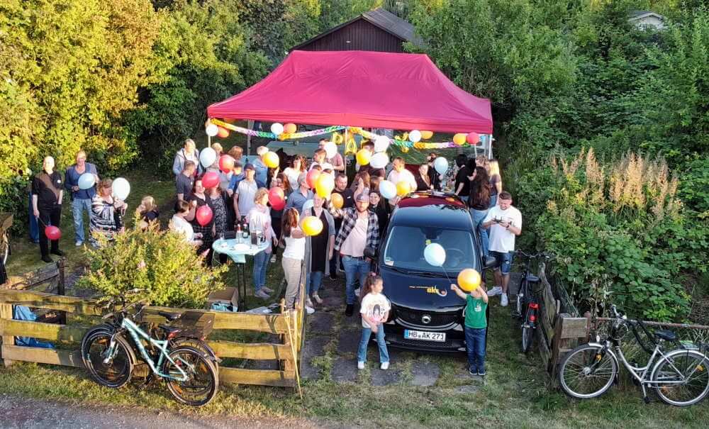 Group of people with balloons under a red gazebo in a garden with trees and a car