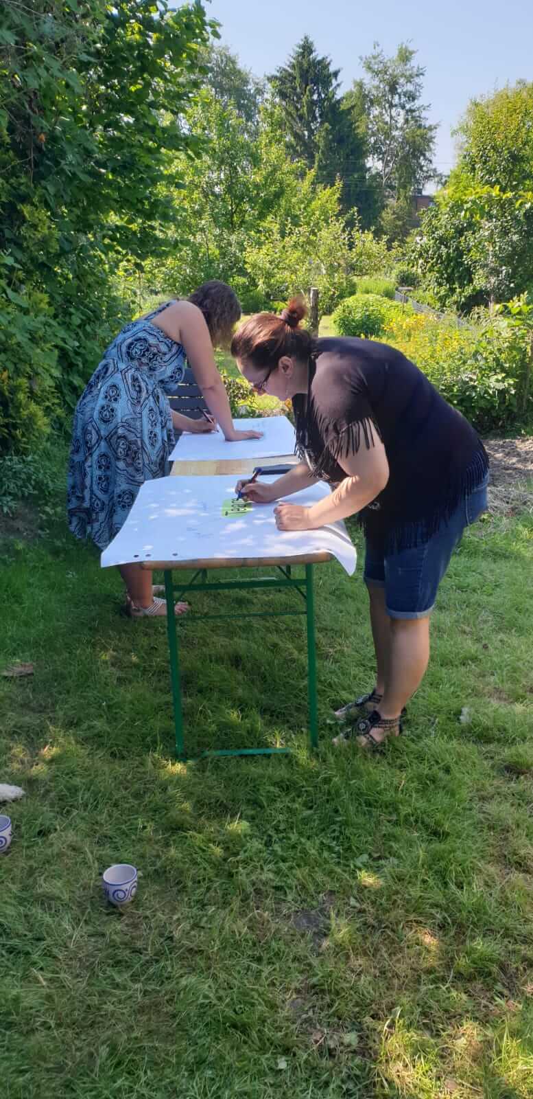 Two women are standing at a white folding table in the garden and writing on paper.