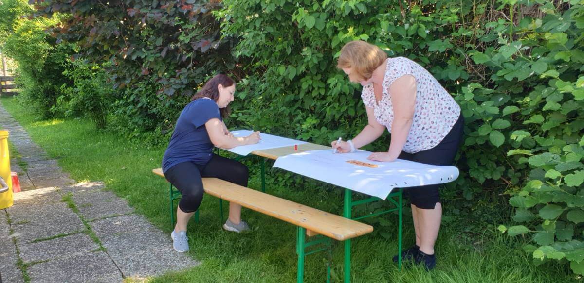 Two women are writing at a long table in the garden, surrounded by plants.