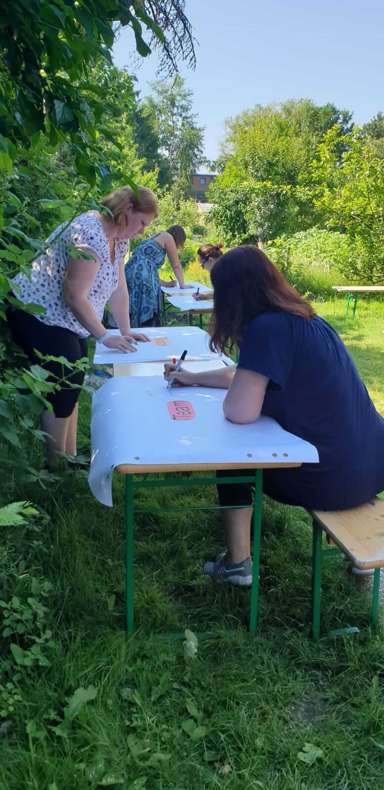 Four women are sitting at a white folding table in the garden and writing on paper.
